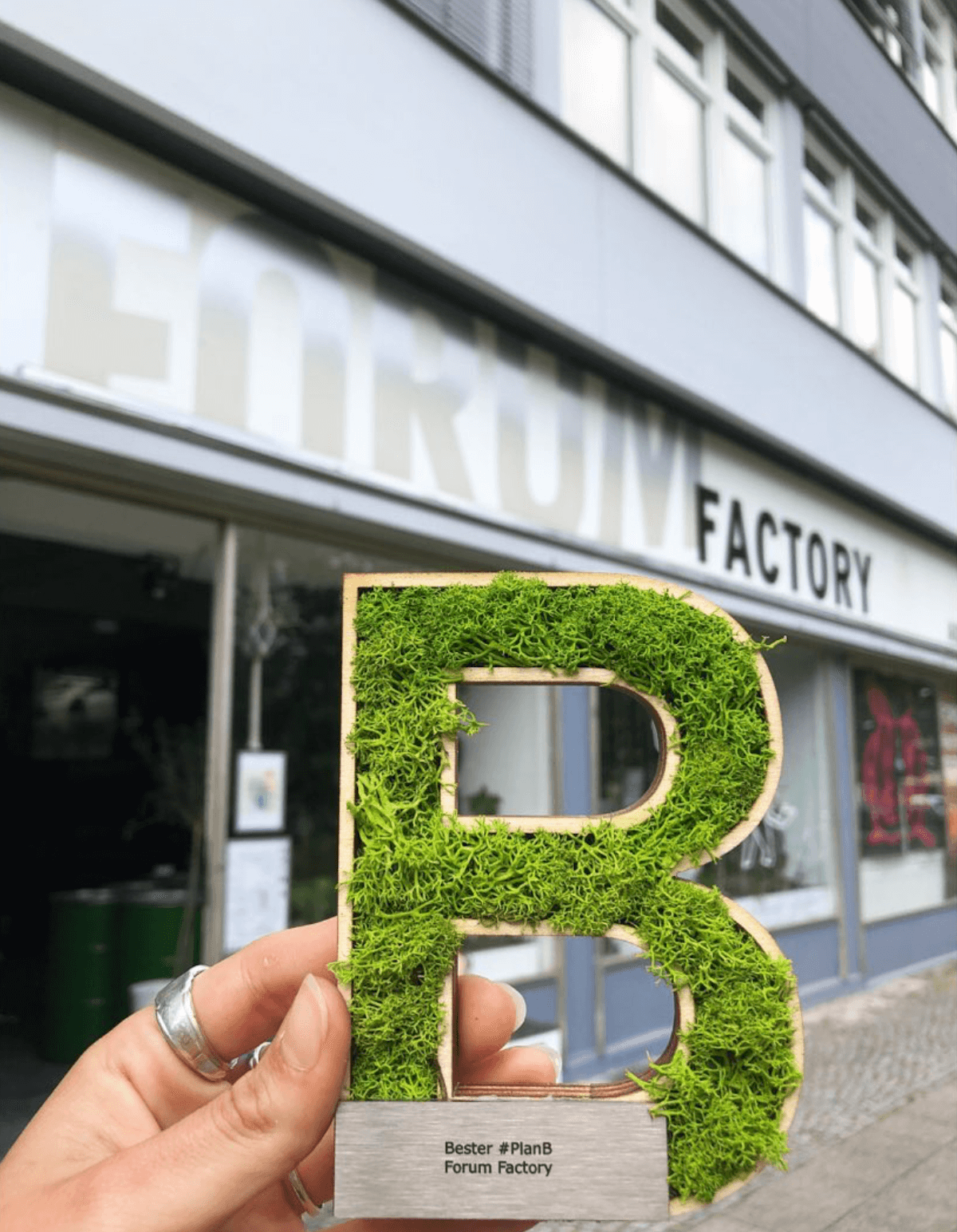 Das Foto zeigt eine Hand, die ein preisgekröntes, mit Moos bedecktes 'B' vor dem Eingang der Forum Factory in Berlin hält. Auf der Metallplakette des Buchstabens steht 'Bester #PlanB Forum Factory'. Im Hintergrund ist der Eingang der Forum Factory mit ihrem markanten Schild zu sehen. Diese nachhaltige Location bietet vielseitige Möglichkeiten für Events und Veranstaltungen